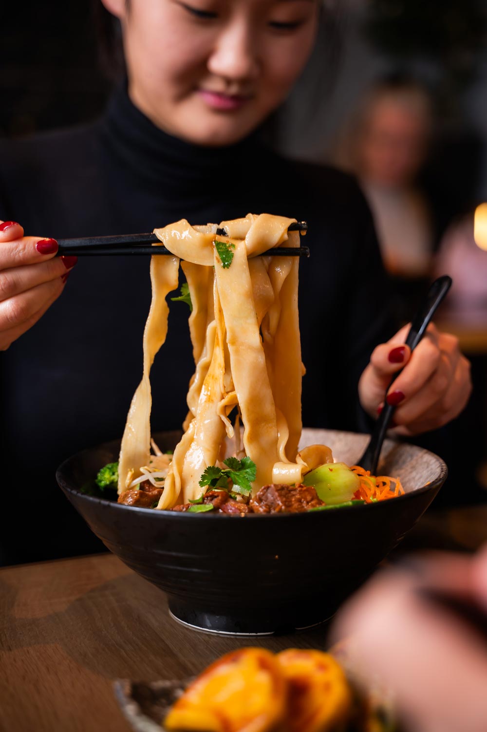 Eine lächelnde Person hält mit Stäbchen breite Nudeln über einer Schüssel.