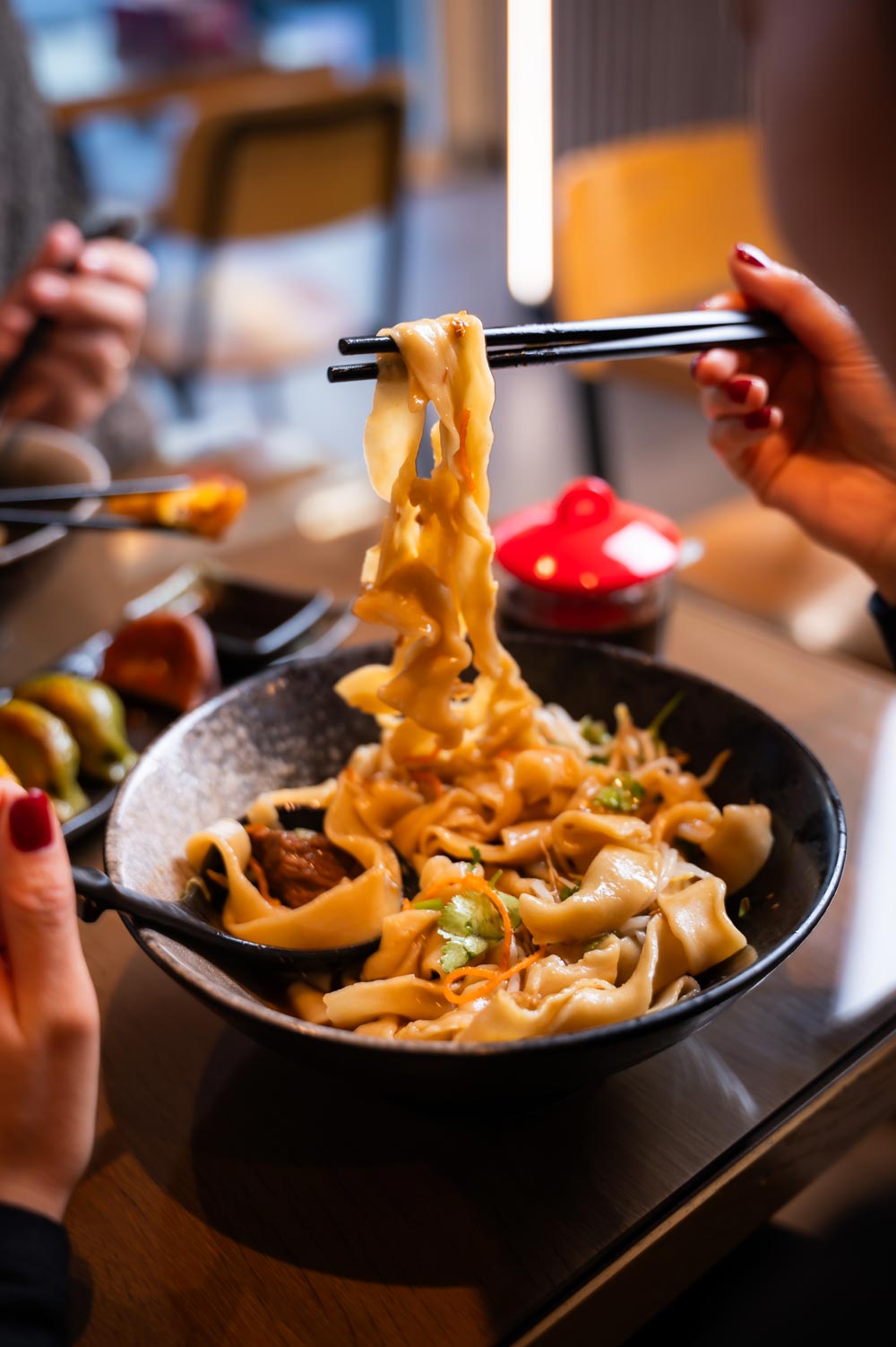 Person is holding chopsticks with noodles over a black bowl filled with dumplings and vegetables, creating a warm and inviting dining scene.