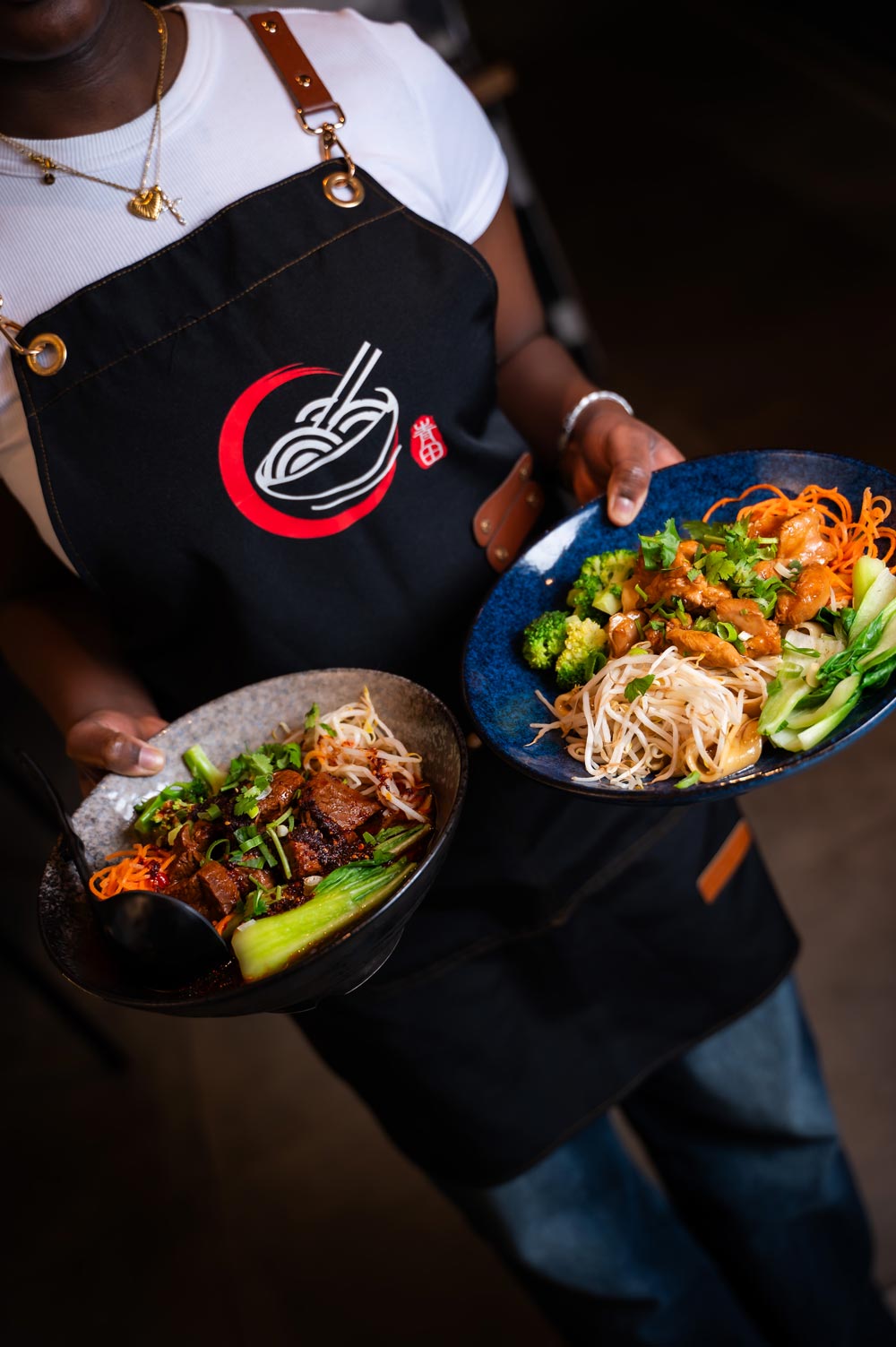 Eine Person trägt eine schwarze Schürze und hält zwei Teller mit asiatischem Essen.